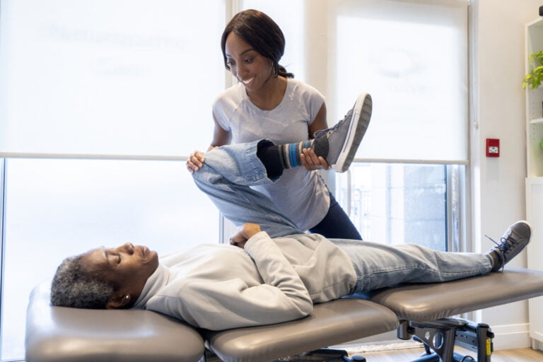 A young female Chiropractor of African decent, works with a senior woman who is laying prone on her treatment table as she evaluates the problem. She is moving the patients leg in various positions and stretches to help pinpoint the trouble area and adjust it accordingly.