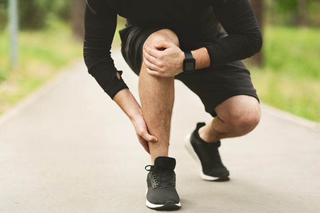 A person in athletic wear is kneeling on a path in a park, holding their lower leg in discomfort. The surrounding environment is green and serene, indicating a daytime workout.